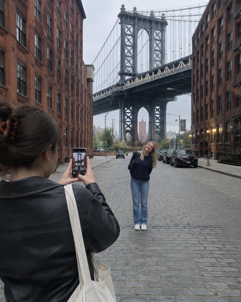 One friend taking an iPhone photo of another on the cobblestones in DUMBO, with the Manhattan Bridge arching behind her
